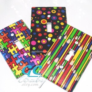 May include: Three colorful light switch covers with different patterns. One cover features a puzzle design with red, yellow, blue, green, and pink pieces. Another cover has a black background with colorful polka dots. The third cover has a red, green, and yellow striped pattern with pencils.