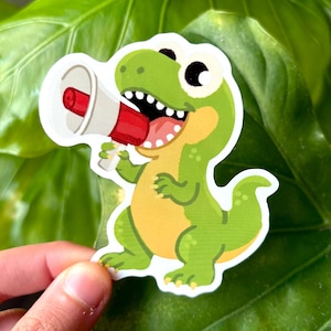 May include: A green dinosaur sticker holding a red and white megaphone. The dinosaur has a yellow belly and is smiling with its mouth open. The sticker is held in front of a large green leaf.