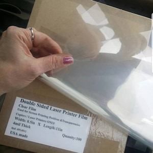 May include: A package of 100 sheets of clear double-sided laser printer film. The film is used for screen printing positives and transparencies. The package is labeled with the text "Double Sided Laser Printer Film", "Clear Film", "Used for Screen Printing Positives & Transparencies", "Copiers/Laser Printers ONLY", "Width: 8.5in X Length:11in", "4mil Thick", "Ref: JPL8511", "USA made", and "Quantity:100".