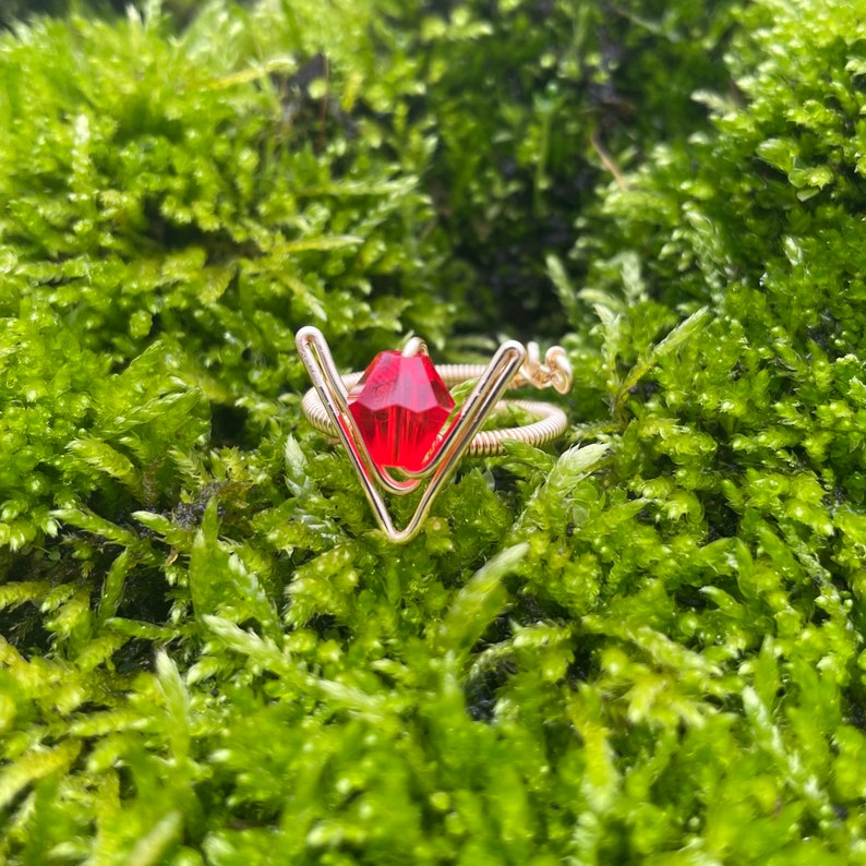 May include: A gold wire ring with a red gemstone in the center. The ring is shaped like a V and is set against a green mossy background.