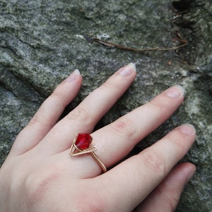 May include: A gold wire ring with a red gemstone. The ring is on a person's finger.