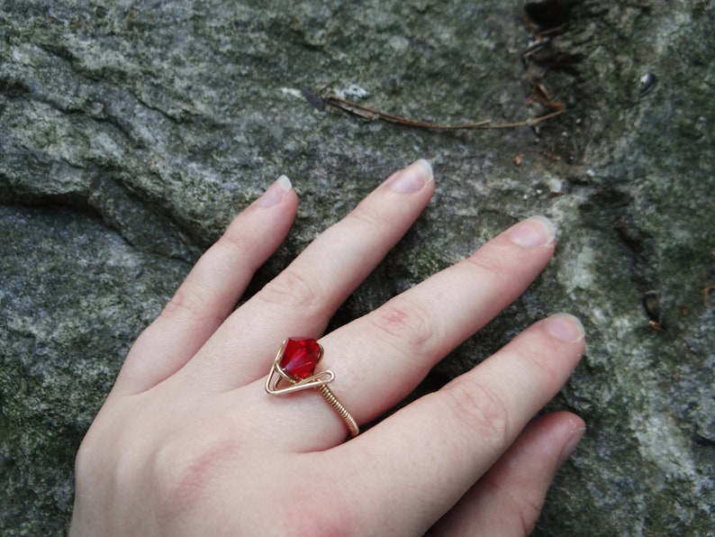 May include: A gold wire ring with a red gemstone. The ring is on a person's finger.