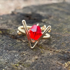 Puede incluir: Un anillo de alambre dorado con una piedra preciosa roja en el centro. El anillo tiene forma de V.