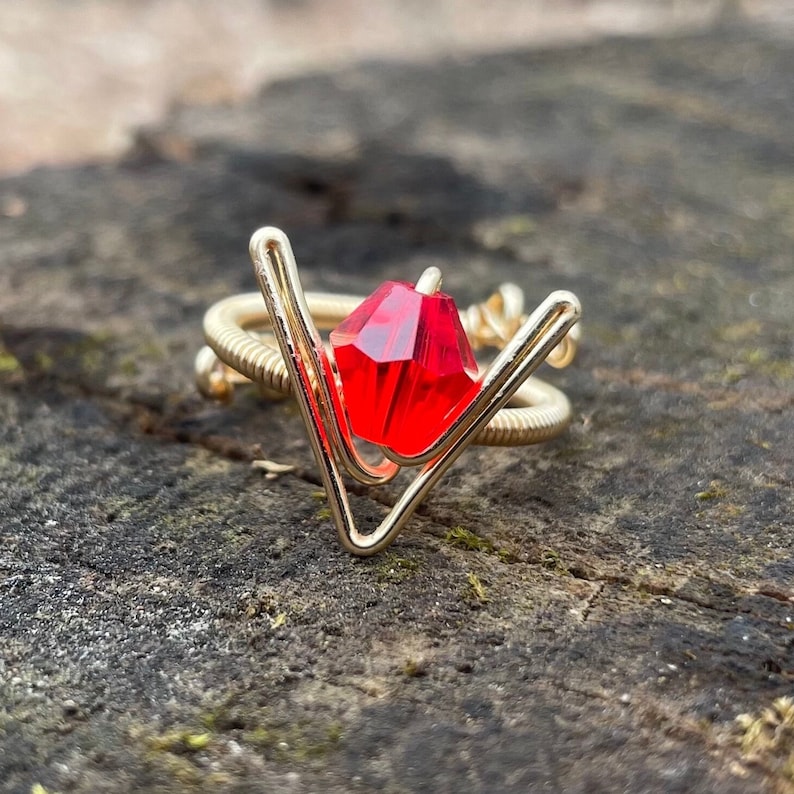 May include: A gold wire ring with a red gemstone in the center. The ring is shaped like a V.