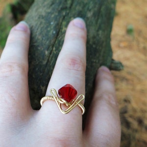 May include: A gold wire ring with a red gemstone. The ring is on a person's finger.