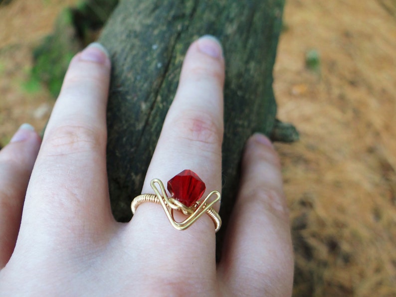 May include: A gold wire ring with a red gemstone. The ring is on a person's finger.