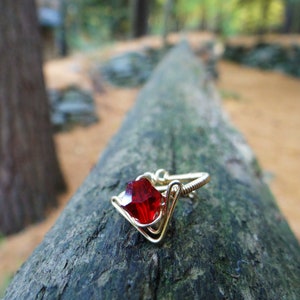 May include: A gold ring with a red gemstone set in a wire-wrapped design. The ring is resting on a tree trunk.