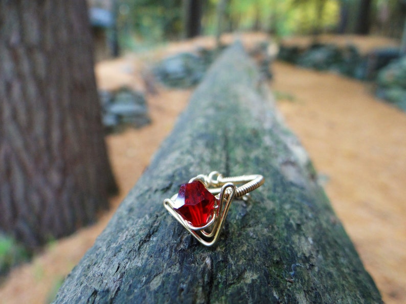 May include: A gold ring with a red gemstone set in a wire-wrapped design. The ring is resting on a tree trunk.