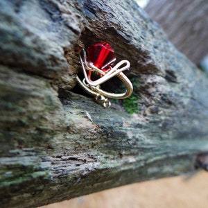 May include: A red gemstone ring made with wire wrapped around a red gem. The ring is sitting on a piece of weathered wood.