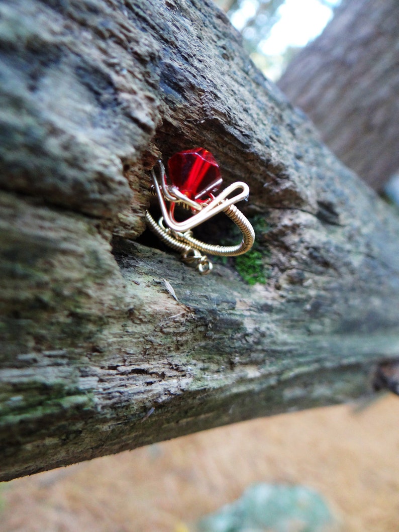 May include: A red gemstone ring made with wire wrapped around a red gem. The ring is sitting on a piece of weathered wood.