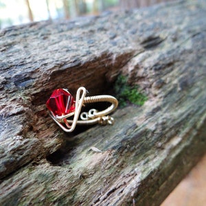May include: A red gemstone ring made with gold wire. The ring is on a wooden surface.