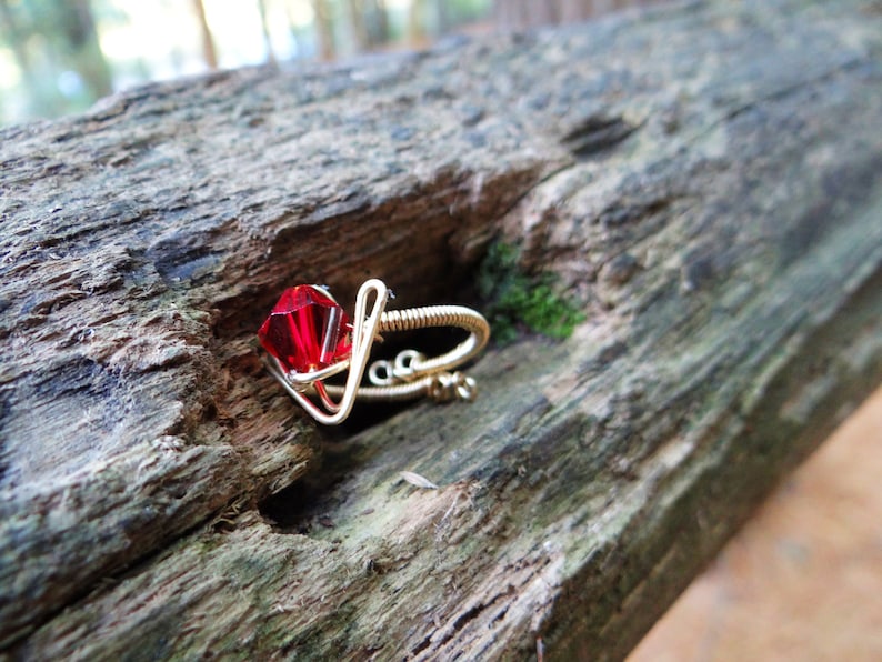 May include: A red gemstone ring made with gold wire. The ring is on a wooden surface.