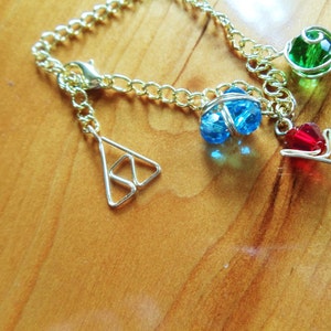 Pode incluir: Um bracelete de corrente dourada com três pingentes: um cristal azul, um cristal verde e uma seta vermelha. O bracelete também tem um pingente em forma de triângulo.