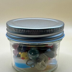 Könnte beinhalten: Ein klares Glas mit silbernem Deckel und blauem Ring, gefüllt mit kleinen, bunten Spielzeugen. Das Glas enthält eine gelbe Quietscheente, einen grünen Oktopus und andere Miniaturgegenstände. Das Glas ist verschlossen.