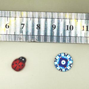 May include: A red ladybug-shaped button and a round button with a blue and white eye design are displayed next to a ruler. The ladybug button is approximately 1.3 cm long. The eye button is approximately 1.3 cm in diameter.
