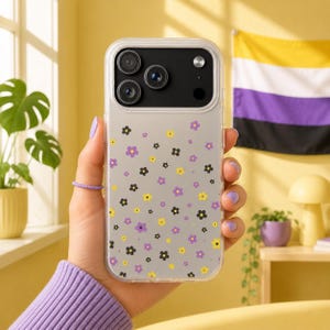 May include: Clear phone case with a floral pattern of purple, yellow, and black flowers. The case is held in a hand with a purple ring and a purple sweater sleeve. The background includes a non-binary pride flag.