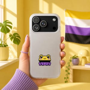 May include: Clear phone case with a cartoon frog sticker in yellow, black, and purple. The frog has a smiling face. The background includes a non-binary pride flag and a yellow wall.