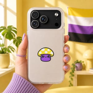 Puede incluir: Funda transparente para teléfono con un diseño de hongo de dibujos animados. El hongo tiene un sombrero amarillo con lunares blancos y un cuerpo morado. La funda del teléfono se sostiene en una mano con esmalte de uñas morado. Una bandera del orgullo no binario está en el fondo.