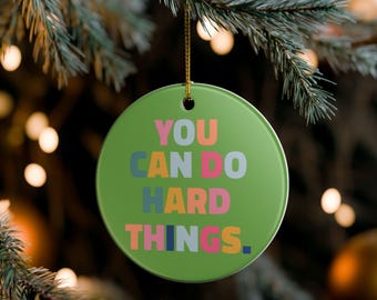 May include: A round, green Christmas ornament with the text "YOU CAN DO HARD THINGS" in colorful block letters. The ornament hangs from a gold string against a blurred background of Christmas tree branches and lights.