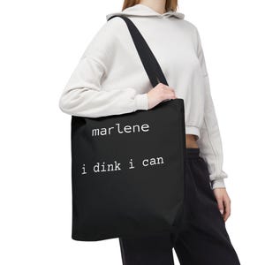 Può includere: Borsa tote nera con il testo bianco "marlene i dink i can". La borsa ha una lunga tracolla nera ed è realizzata in materiale resistente. Questa borsa tote è progettata per trasportare oggetti ed è adatta all'uso quotidiano.