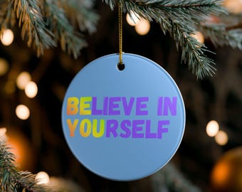 May include: A light blue, round Christmas ornament with the words "BELIEVE IN YOURSELF" in colorful gradient text. The ornament hangs from a gold string against a blurred background of Christmas tree branches and lights.
