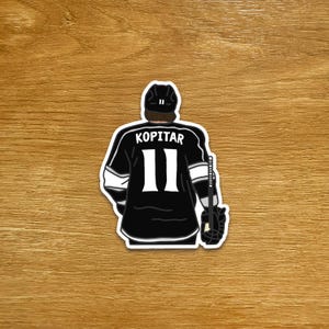 Peut inclure: Un autocollant représentant le maillot d'un joueur de hockey de dos. Le maillot noir a des rayures blanches et le nom "KOPITAR" au-dessus du numéro "11". Le joueur porte un casque noir et tient une crosse et un gant de hockey.