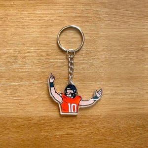 May include: A clear acrylic keyring featuring a football player in an orange jersey with the number 10. The player is wearing a black helmet and has his arms raised in a celebratory gesture. The keyring has a silver metal ring and chain.