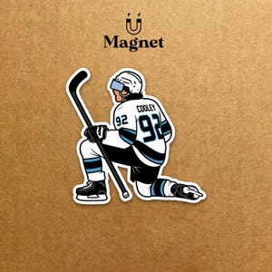 May include: A sticker of a hockey player kneeling, holding a hockey stick. The player wears a white jersey with black and light blue accents, the number 92, and the name "Cooley". The sticker has the word "Magnet" at the top.