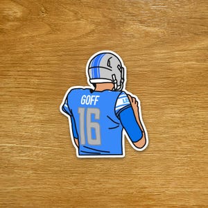 May include: A sticker depicting a cartoon football player in a blue jersey with the name "GOFF" and the number "16". The player wears a silver helmet with blue stripes. The sticker has a white border, and is set against a wooden background.