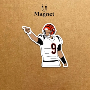 May include: A white magnet featuring a cartoon illustration of a football player in a white jersey with the number 9 and a helmet with orange and black stripes. The player is pointing with his right hand. The word "Magnet" is above the image.