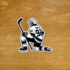 May include: A sticker depicting a hockey player kneeling, wearing a white and black jersey with the number 92. The player holds a hockey stick. The sticker has a white border and is on a wooden surface.