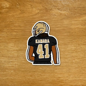 Peut inclure: Autocollant représentant un joueur de football américain en uniforme noir et or. Le maillot du joueur porte le nom "KAMARA" et le numéro "41". Le joueur porte un casque doré. L'autocollant est sur une surface en bois.