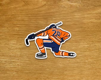 May include: A sticker featuring a hockey player in an orange and blue uniform, kneeling on one knee. The player is holding a hockey stick. The jersey number is 20. The sticker is on a wooden surface.