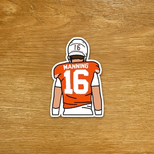 Op de afbeelding: Een sticker met een American footballspeler in een oranje shirt met nummer 16 en de naam MANNING. De speler is van achteren te zien, met een witte helm met nummer 16.