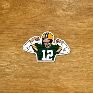 May include: A sticker featuring a cartoon illustration of a football player flexing his arms. The player wears a green and yellow uniform with the number 12. The helmet is yellow with a green stripe.