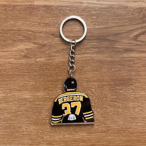 May include: A hockey jersey design keychain. The black jersey has yellow stripes, the name "BERGERON" and the number "37" in yellow. The keychain has a silver ring and chain.