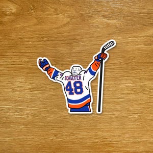 May include: A sticker depicting a hockey player in a white, blue, and orange jersey with the number 48 and the name "Schaefer." The player is raising a hockey stick. The sticker is on a wooden surface.