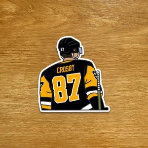 May include: A sticker depicting a hockey player from the rear, wearing a black and gold jersey with the number 87 and the name "CROSBY". The player is wearing a black helmet with a visor and holding a hockey stick.