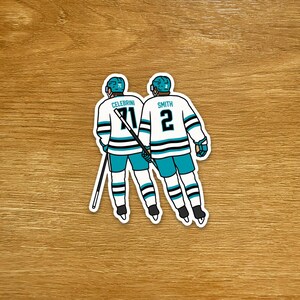 Può includere: Un adesivo con due giocatori di hockey visti da dietro, con maglie turchesi e bianche. Una maglia ha il nome "Celerbrini" e il numero "71", l'altra "Smith" e il numero "2". L'adesivo è su una superficie di legno.