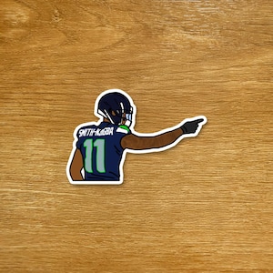 Op de afbeelding: Een sticker met een cartoon illustratie van een American football speler in een marineblauw shirt met nummer 11 en de naam "Smith-Njigba". De speler wijst met zijn rechterarm. De sticker heeft een witte rand.