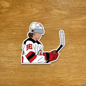 Può includere: Un adesivo raffigurante un giocatore di hockey su ghiaccio con una maglia bianca con dettagli rossi e neri. Il giocatore indossa un casco e guanti rossi, tenendo una mazza da hockey. La maglia mostra il numero 86.