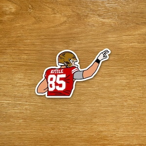 May include: A sticker depicting an American football player in a red jersey with the number 85. The player wears a gold helmet and gestures with his right hand. The name "Kittle" is above the number. The sticker is on a wooden surface.