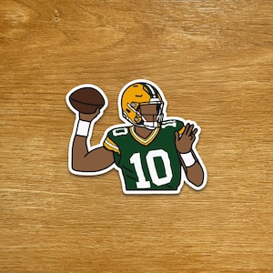 May include: A sticker featuring an American football player in a green and yellow uniform, throwing a brown football. The player wears a helmet with a yellow face mask and the number 10 on the jersey.