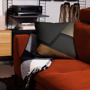 Könnte beinhalten: Ein orangefarbenes Sofa mit einem geometrisch gemusterten Kissen in Schwarz, Braun und Gold. Eine schwarz-weiß gefranste Decke ist über das Sofa drapiert. Eine schwarze Stereoanlage steht auf einem Regal und schafft einen modernen Wohnraum.