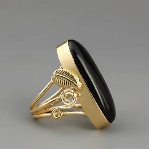 May include: A gold-toned ring featuring a large, smooth, elongated oval black stone set vertically. The band is crafted with decorative metalwork, including a small leaf-shaped element and delicate swirling wire details on the side of the setting.