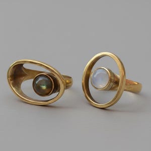 May include: Two gold-toned rings featuring open oval bands. The left ring holds a dark, iridescent round stone, while the right ring features a smooth, translucent white round stone centered within the open metal frame of the band.