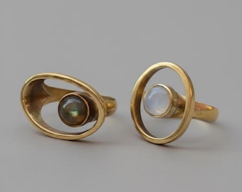May include: Two gold-toned rings featuring open oval bands. The left ring holds a dark, iridescent round stone, while the right ring features a smooth, translucent white round stone centered within the open metal frame of the band.