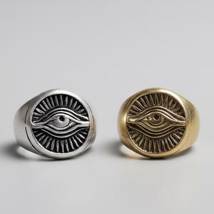 May include: Two signet rings, one silver and one gold, each featuring an eye design. The rings have a circular face with an eye symbol at the centre, surrounded by radiating lines. The background is plain.