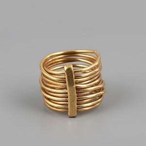 May include: A gold-colored ring with multiple coiled bands held together by a vertical bar. The ring has a stacked design, with each band visible. The ring is set against a gray background.