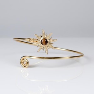 May include: Gold-toned arm cuff bracelet featuring a sunburst design with a brown gemstone center. The bracelet has a spiral detail at one end and a decorative clasp. A stylish piece of jewelry.